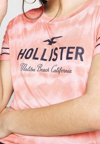 Pink tie-dye t-shirt featuring black "Hollister" logo and "Malibu Beach California" text. Short sleeves with black stripes on shoulders.