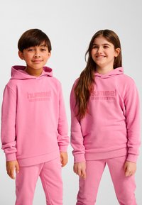 Zwei Kinder tragen passende pinke Kapuzenpullover und Hosen aus weichem Stoff mit einem strukturierten Logo auf der Brust.