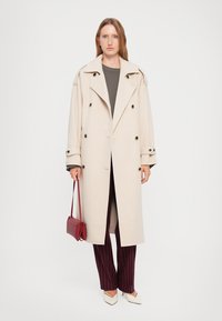 Beige Doppelreiher-Mantel mit schwarzen Knöpfen, großem Kragen, kombiniert mit einem dunklen Oberteil und bordeauxfarbenen gestreiften Hosen, hält eine kleine rote Tasche.