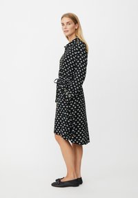 Czarna sukienka z białym geometrycznym wzorem, z kołnierzykiem, długimi rękawami i wiązaniem w talii. Luźna tkanina, długość do kolan, noszona z czarnymi balerinami.