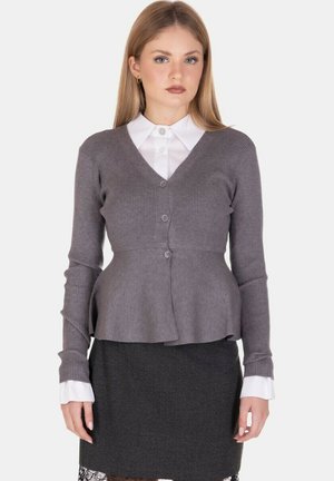 Cardigan grigio a coste con scollo a V, caratterizzato da un busto aderente e un orlo svasato, abbinato a una camicia bianca con colletto e una gonna scura.
