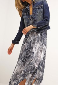 Tummansinisestä denim-takista, jossa on napit, on yhdistetty sinisen ja harmaan sävyissä kuvioituun maksimekkoon, jossa on korkea halkio.