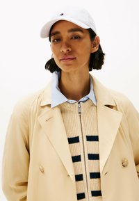 Beige trenchcoat over een crème en marine gestreepte gebreide trui, aangevuld met een lichtblauwe overhemde en een witte baseballpet.