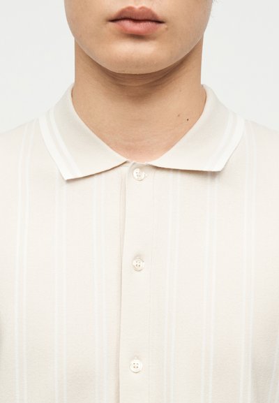 Κοντινό πλάνο ενός ατόμου που φοράει ένα μπεζ πόλο με κουμπιά, διακριτικές κάθετες ρίγες και κλασικό γιακά.