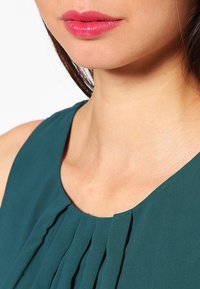 Robe sans manches turquoise avec un col plissé, mettant en valeur un tissu lisse et léger. Mise au point sur la clavicule et les lèvres avec une teinte rose.