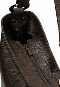 Close-up van een open, donkerbruin, textuurleer tas met een centrale zwarte rits en een gedeeltelijk zichtbare schouderriem, bevestigd met klinknagels.