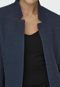 Blazer a scacchi blu navy con colletto alto e apertura frontale, abbinato a un top a canotta nero a coste sotto.