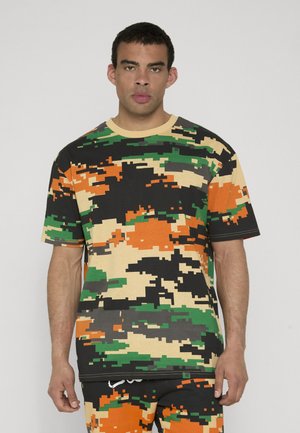 Mann iført kortermet skjorte og shorts med digitalt kamuflasjemønster i grønt, oransje, svart og beige, står mot en ensfarget bakgrunn.