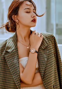 Reloj de pulsera negro con esfera redonda y acentos de strass, combinado con un collar de colgante dorado, usado con un blazer de cuadros sobre una blusa crema.