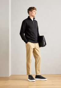 Suéter negro de cuello con cremallera, chinos beige y zapatillas negras con suelas blancas. Modelo sosteniendo un bolso mensajero negro y estructurado. Ambientación minimalista.