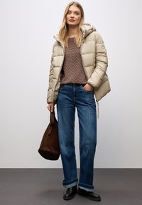 Giubbotto imbottito beige con cappuccio, maglione texture marrone, jeans blu e stivali neri alla caviglia, accessorati con una borsa a tracolla in suede marrone.