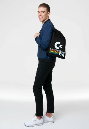Schwarzer Rucksack mit Kordelzug, der das Logo "Commodore 64" in Weiß und gestreiften Akzentfarben zeigt. Das Modell trägt eine blaue Bomberjacke und schwarze Hosen.