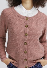 Cardigan en maille de couleur rose avec un motif texturé, doté d'un col rond, de poignets côtelés et de grands boutons marron sur le devant.