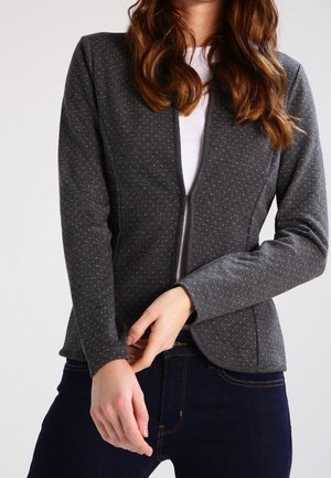 Woman wearing a fitted dark gray blazer with subtle dot pattern over a white top and dark blue jeans, posed with hands clasped.