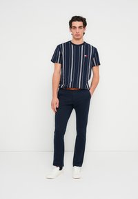 Μπλε σκούρο κοντομάνικο t-shirt με λευκές κάθετες ρίγες, συνδυασμένο με μπλε σκούρο παντελόνι και λευκά αθλητικά παπούτσια. Η καφέ ζώνη προσθέτει λεπτομέρεια.