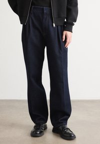 Person iført løse, mørkeblå jeans, sorte lædersko og en sort lynlåsjakke, der står på et glat gråt gulv.