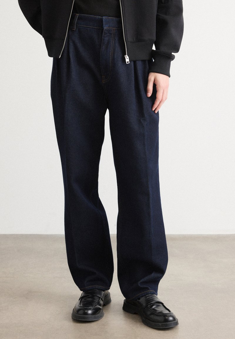 Person iført løse, mørkeblå jeans, sorte lædersko og en sort lynlåsjakke, der står på et glat gråt gulv.