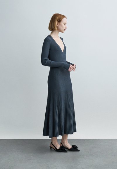 Γυναίκα με κοντά κόκκινα μαλλιά που φοράει μακρύ, σκούρο ριμπ φόρεμα με βαθύ λαιμόκοψη σε σχήμα V και μαύρες γόβες με λεπτομέρειες λουλουδιών, όρθια πλευρικά.