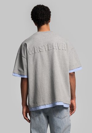 T-shirt oversize gris avec "WRSTBHV" brodé au dos, présentant des accents rayés bleus aux manches et à l'ourlet.