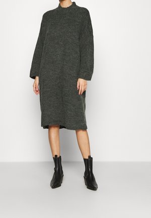 Vrouw die een donkergrijs lang gebreide jurk met lange mouwen en zwarte enkellaarzen draagt, staand tegen een eenvoudige lichte achtergrond.