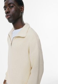 Cremfarbener Strickpullover mit Viertelreißverschluss-Kragen, gerippter Textur und fallenden Schultern. Über einem weißen Shirt getragen, das einen lockeren Schnitt hat.