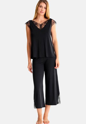 Femme portant un haut noir sans manches et un pantalon large orné de dentelle, debout pieds nus devant un fond blanc.