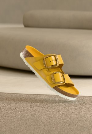 Gele open-teen sandaal met twee verstelbare gouden gespen, kurken voetbed en witte zool, geplaatst tegen een beige bank als achtergrond.