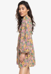 Vestido mini floral con patrones multicolores de flores amarillas, rosas y verdes sobre un fondo rosa claro; con mangas largas y abullonadas.