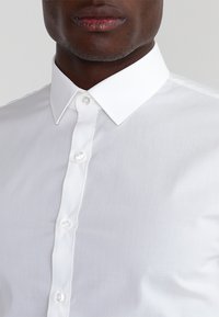 Vit skjorta med knappar framtill, klassisk krage, slät textur och rundvita knappar på knappraden. Närbild visar halsringning och tyg.