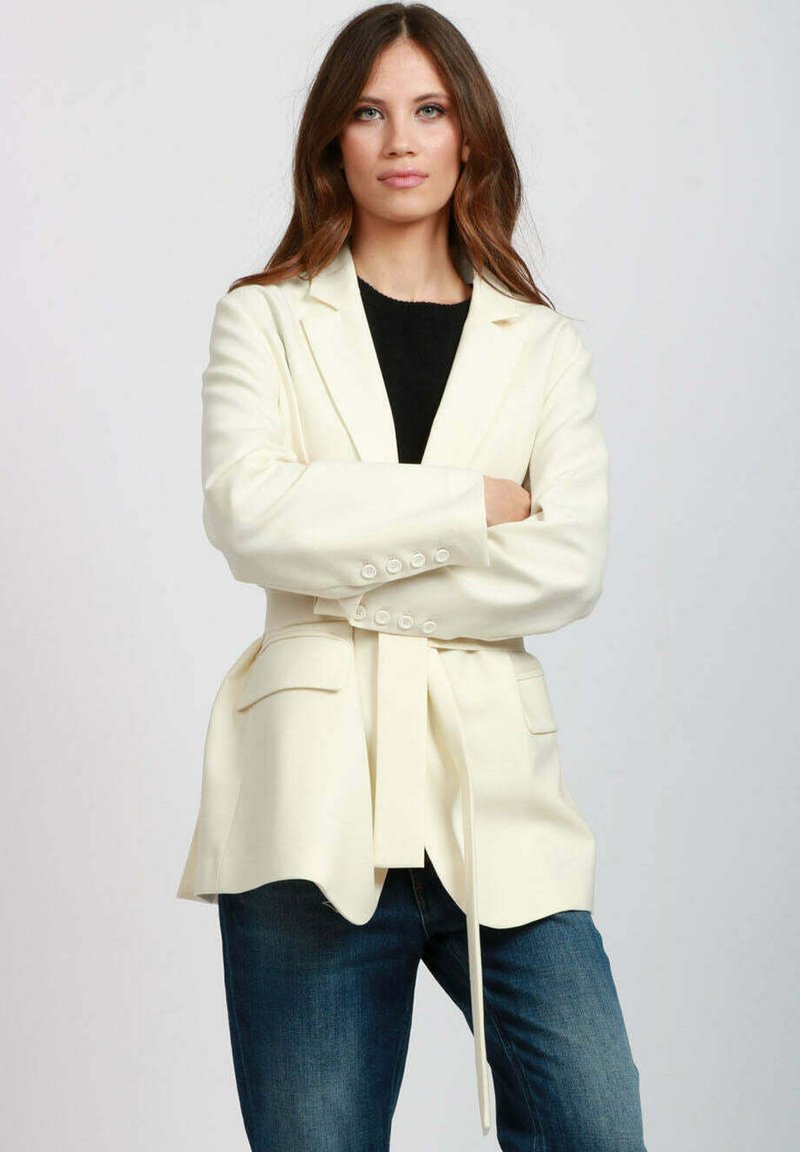 Blazer color crema con una vestibilità strutturata, ampi rever, chiusura a doppio bottone, cintura in vita e due tasche frontali. Indossato sopra un top nero.