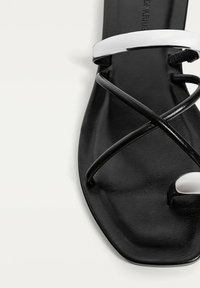 Svart platt sandal med rektangulär tåform, med glansiga vita och svarta remmar som korsar ovanpå. Tillverkad av syntetiskt material.