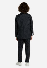 Completo nero a righe con giacca strutturata e pantaloni a gamba dritta. Sneakers bianche visibili. Cuciture curate e dettagli con bottoni sulle maniche.