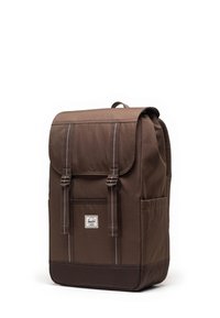 Sac à dos en toile marron avec un rabat, deux sangles avant, un grand compartiment principal et des poches latérales. Présente un patch logo.