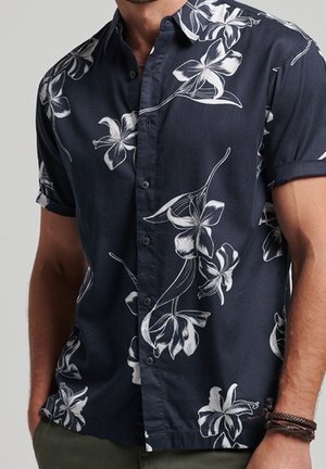 Man wearing a short-sleeve navy shirt with white floral prints, partially unbuttoned, and a braided leather bracelet on left wrist.