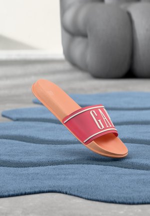 Sandale à glissière rouge et pêche avec logo "GAP" sur la sangle, reposant sur un tapis bleu texturé avec un siège rembourré gris en arrière-plan.