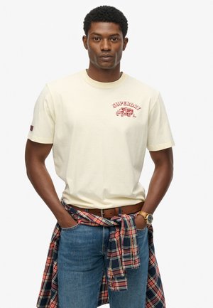 Cream short-sleeve t-shirt with red "SUPERDRY" graphic and truck design. Worn with blue jeans and a plaid shirt tied at the waist.