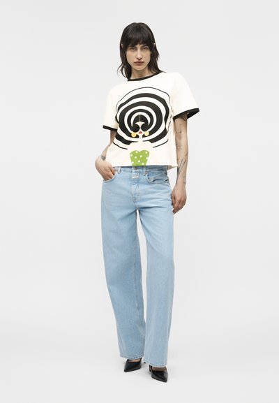 Femeie cu păr negru, purtând un tricou alb cu un design spiralat alb-negru și detalii cu buline verzi, blugi albaștri deschiși și pantofi cu toc negri.