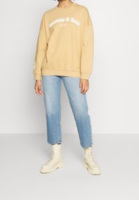 Sudadera amarilla con el texto "Sunshine & Rain", combinada con jeans azul claro y botas beige con cordones; ajuste relajado y estilo casual.