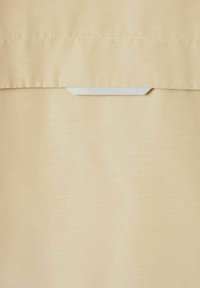 Tejido beige con una pequeña etiqueta triangular blanca cosida debajo de una costura horizontal, sugiriendo un detalle de cierre o sujeción.