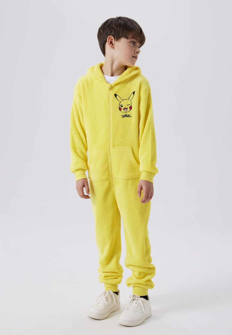 Gelbes Fleece-Overall mit Kapuze und Fronttaschen, mit einem gestickten Pikachu-Design auf der Brust. Kombiniert mit weißen Sneakers.