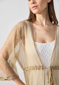 Duster dorado transparente con brillo metálico. Cuenta con una cintura atada y un frente abierto, con suaves pliegues en una prenda interior blanca. Manga corta.