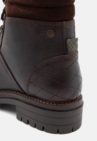 Barna bőr csizma strukturált felülettel, steppelt részletekkel és sötétbarna gumi talppal. Hátsó részén logóval és csíkos szövet díszítéssel rendelkezik.