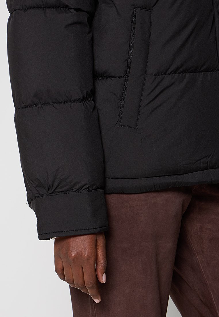 La chaqueta acolchada negra presenta un diseño acolchado con un grosor de aislamiento. La manga tiene un acabado de puño, y hay un bolsillo lateral para almacenamiento.