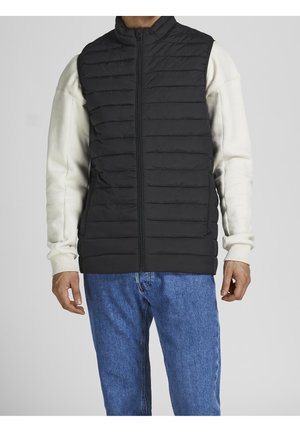 Schwarze gepolsterte Weste mit hohem Kragen und gesteppter Textur, getragen über einem weißen Sweatshirt, kombiniert mit blauen Jeans. Mit Reißverschluss vorne.