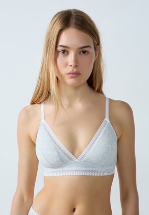 Giovane donna con lunghi capelli biondi che indossa un reggiseno grigio chiaro con coppe morbide e rifiniture in pizzo bianco su uno sfondo chiaro uniforme.