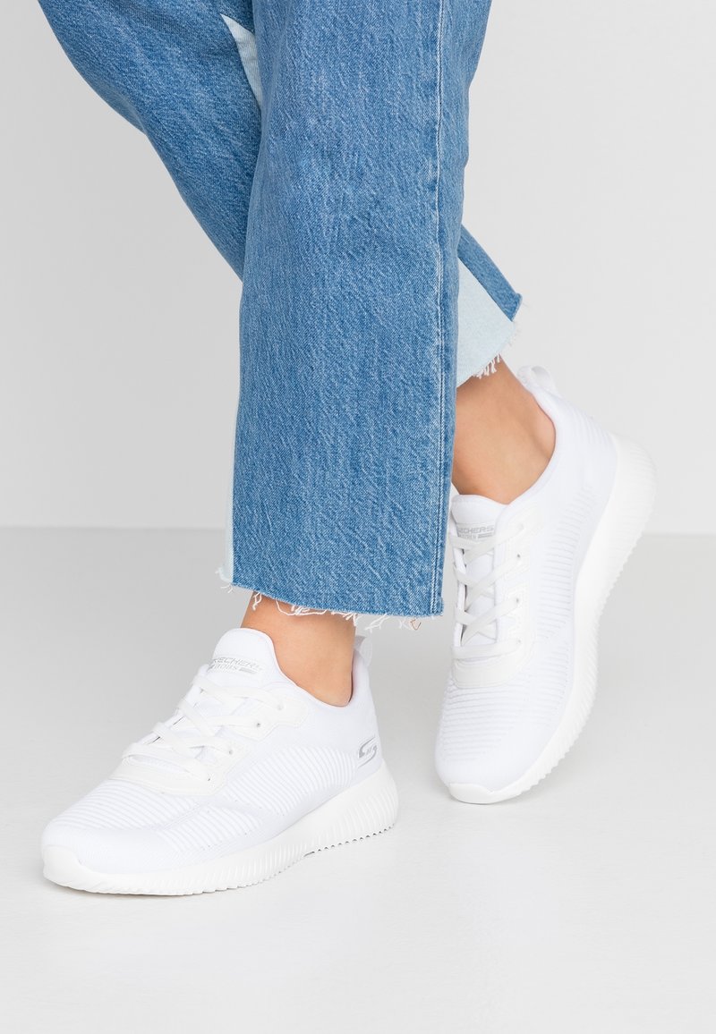 Vita sportskor med ett strukturerat textilt ovandel och gummisula, med snörning och logotyp på sidan, i kombination med korta blå jeans.