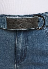 Jeansbälte i denim med ett svart texturerat tygband över en silverfärgad metalld-ring, fäst vid blå denimjeans med synlig ficksyddning.