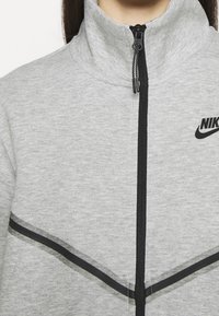 Chaqueta gris con cremallera y cuello alto. Presenta una cremallera negra, rayas negras en el pecho y un logo de Nike en el lado derecho. Textura suave.