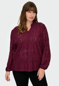 Bordo bluzica z zapletenimi čipkastimi detajli, dolgimi napihnjenimi rokavi, V-izrezom in sproščenim krojem. V kombinaciji s črnimi hlačami.