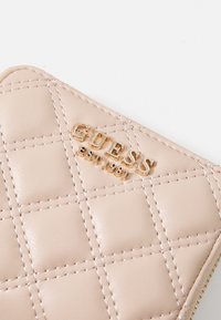 Quiltet beige faux lærred lommebok med gull "GUESS" logo, avrundede hjørner og glidelås i toppen med kontraststingdetaljer.